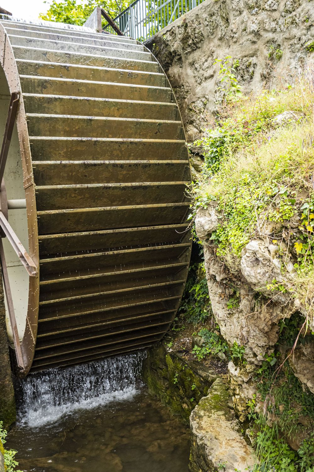 Close-up view of a historic wooden water wheel turning in a stone mill race with cascading water and green foliage.