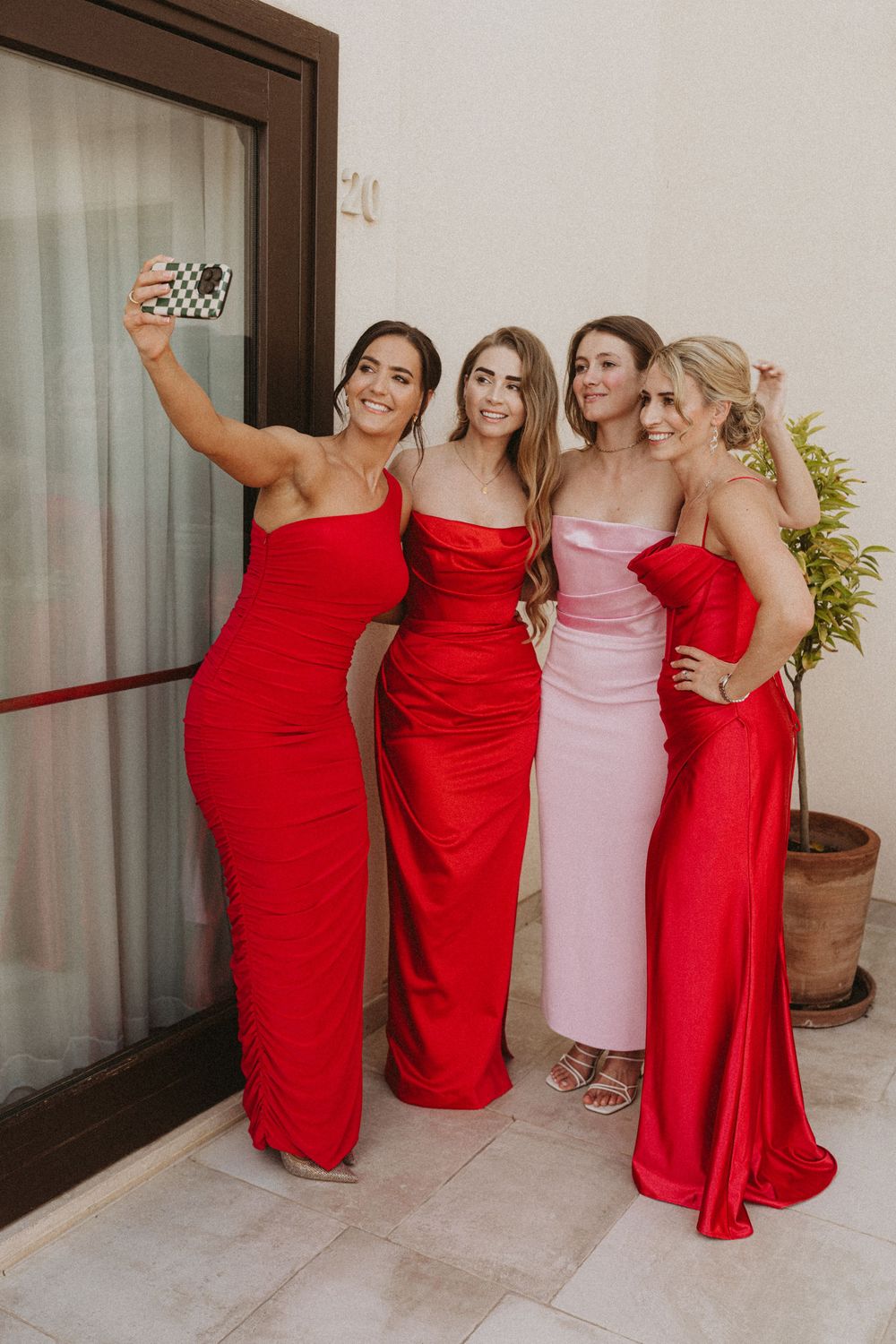 A group of bridesmaids in red and pink formal dresses pose together for a selfie at a wedding.