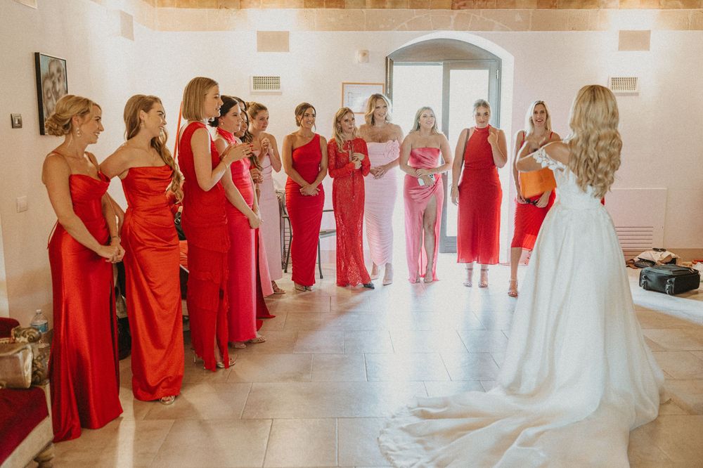 Bridesmaids in matching red dresses gather in a bright room with the bride wearing a flowing white gown.