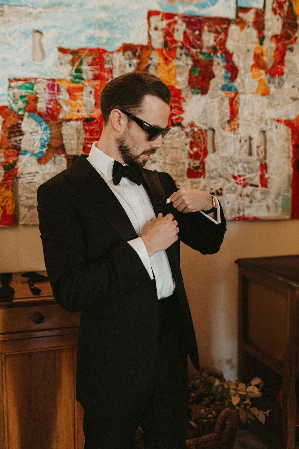 Two people in formal black tuxedos sharing a drink together against a colorful wall with artwork.