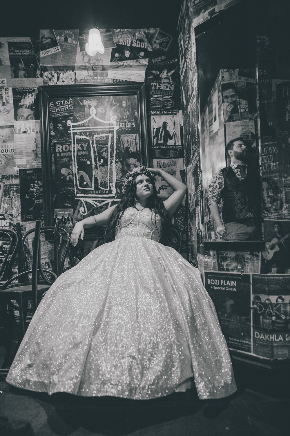 A person in a sparkling ballgown poses against a wall covered in vintage posters and signs in black and white photography.