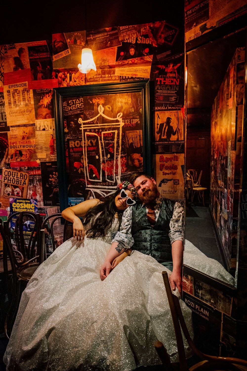 A bride in a white gown lies across a vintage wall covered in rock music posters and dim lighting.