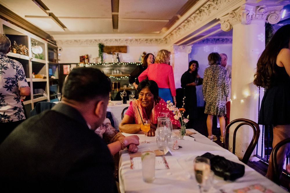 Wedding guests enjoy an elegant reception dinner in an ornate ballroom with decorative crown molding and purple lighting.