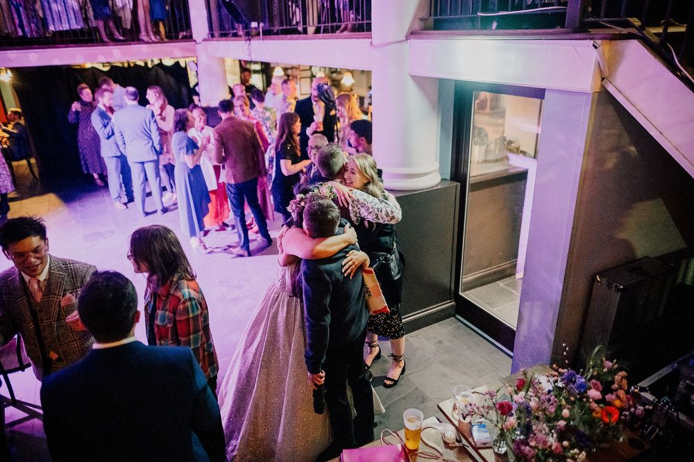 Wedding guests dance and celebrate on a crowded dance floor illuminated by purple lighting at an indoor reception venue.
