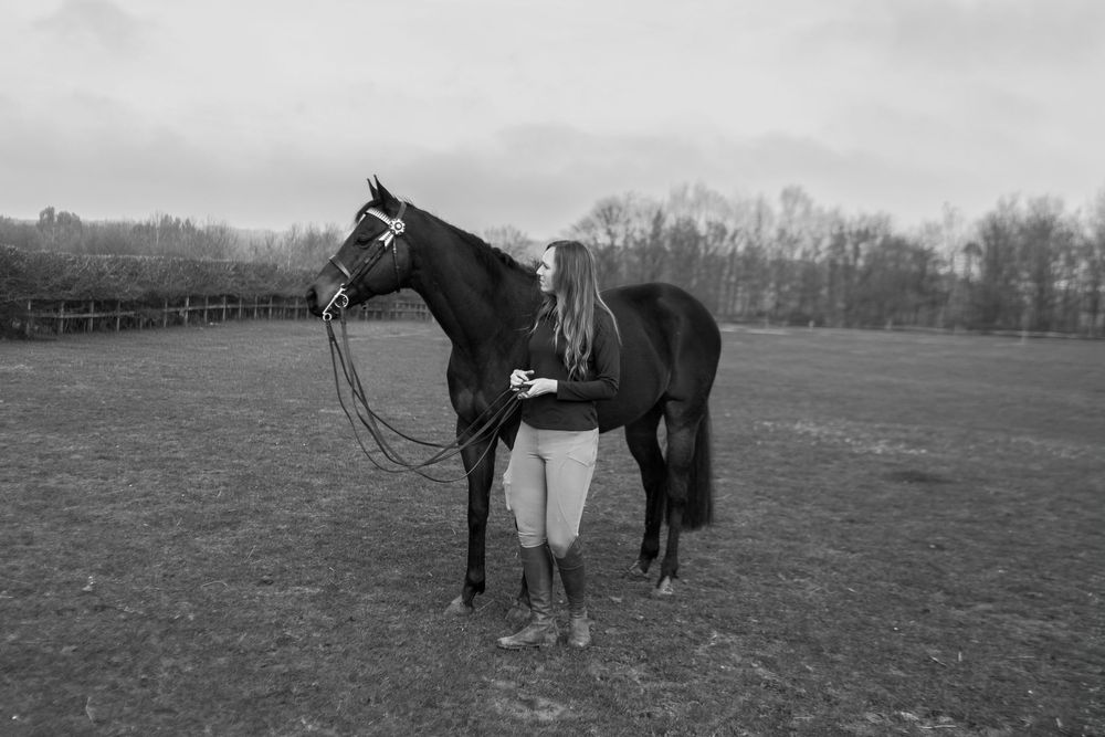 Black and white photo of a young woman standing with a dark horse in an open field with trees in the background. Essex Equine Photographer.