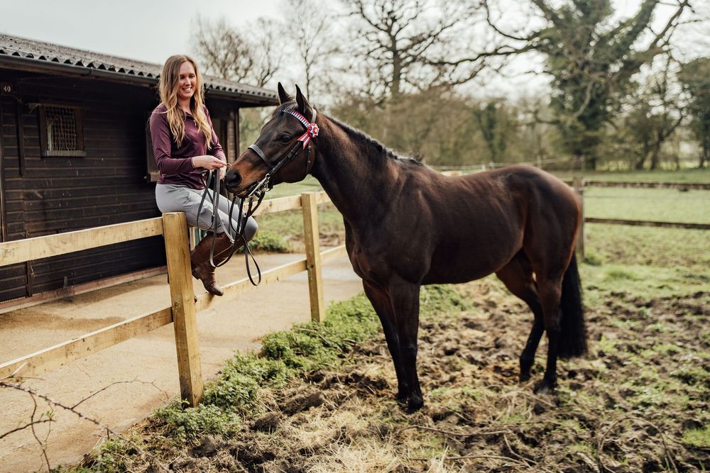 Young woman standing on fence beside a dark brown horse at a rural stable yard on a cloudy day. Essex Equine Photographer.
