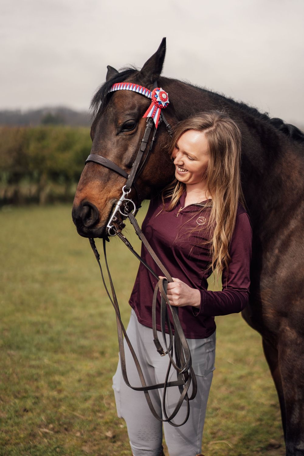 Woman in burgundy shirt smiling beside a bay horse wearing a rosette ribbon in a green field. Essex Equine Photographer.
