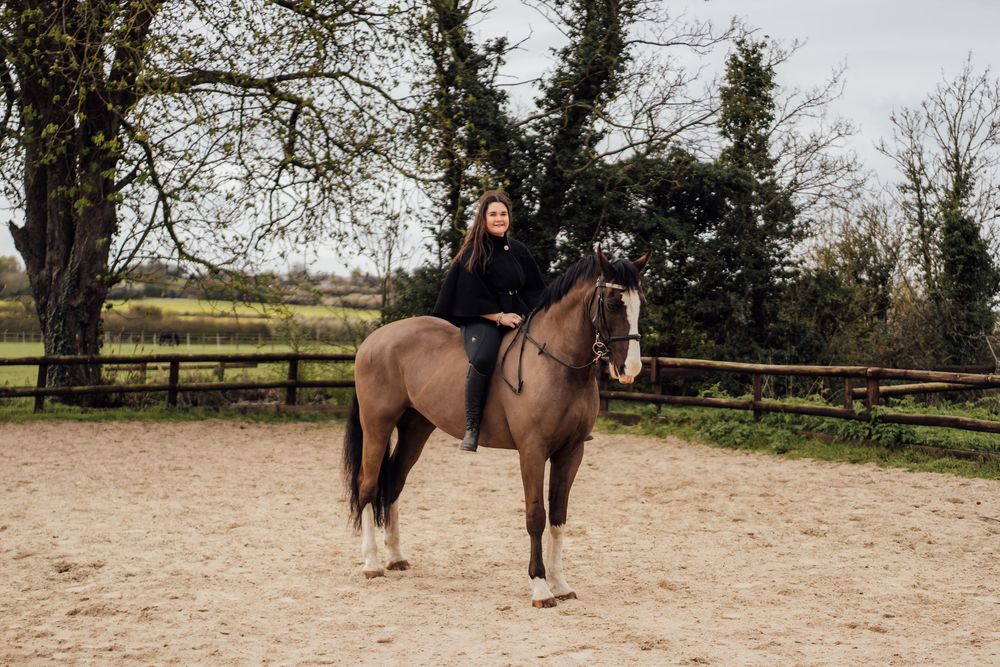 A rider in black attire sits atop a brown horse in a sandy equestrian arena surrounded by trees. Essex Equine Photographer.