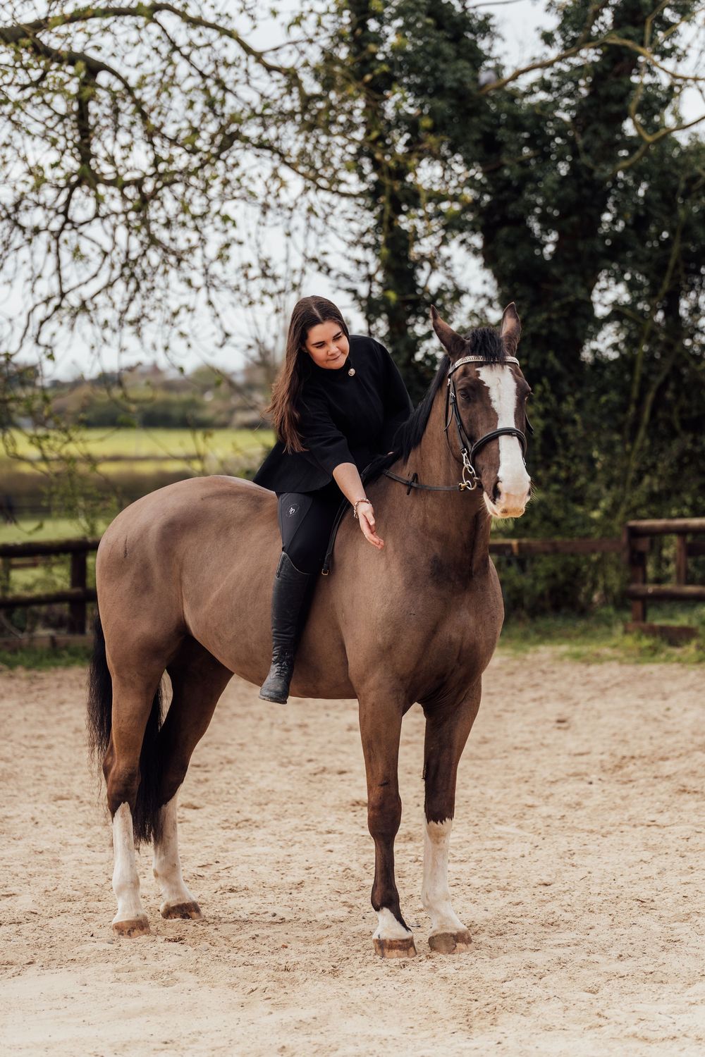 Young woman in black outfit smiling while riding a dapple grey horse in an outdoor sand arena. Essex Equine Photographer.