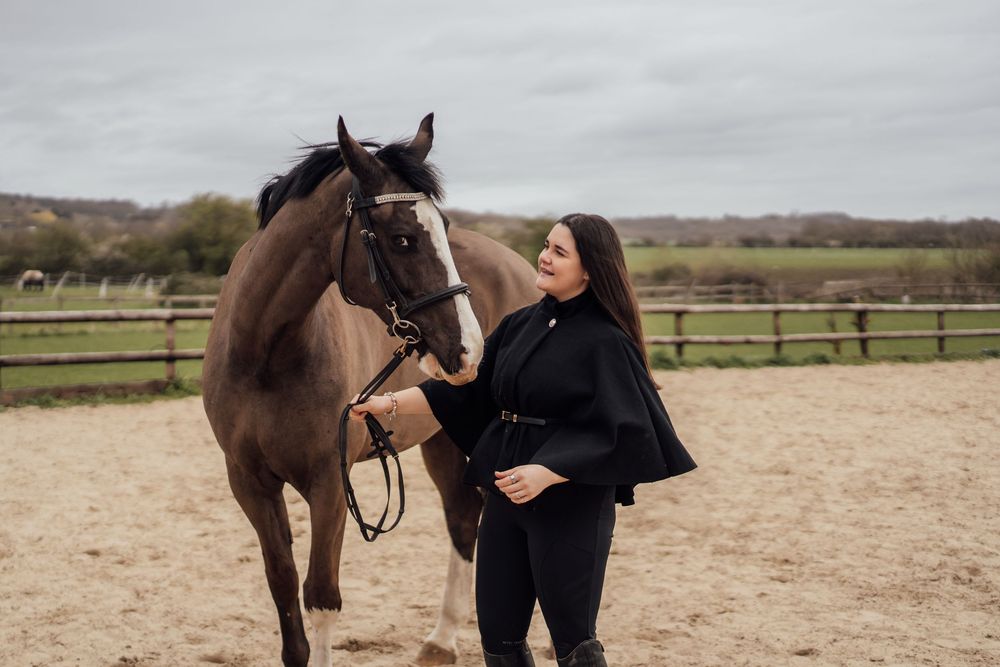 Pregnant woman in black outfit standing beside a brown horse in a sandy equestrian arena on a cloudy day. Essex Equine Photographer.