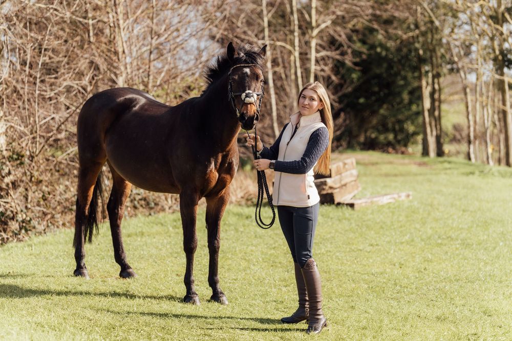 Woman in equestrian attire standing beside a dark bay horse on green grass with trees in background. Essex Equine Photographer