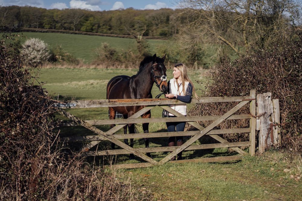 Woman in white jacket bonding with a dark horse over a rustic wooden gate in a lush green countryside.  Essex Equine Photography.