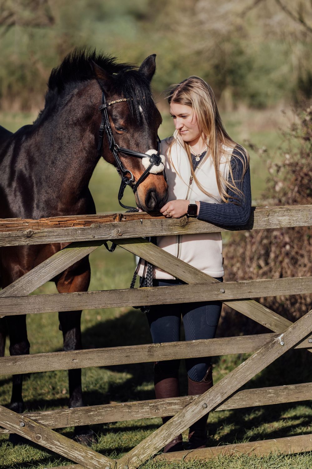 Blonde woman in a white vest tenderly interacts with a dark bay horse over a wooden gate in a sunny paddock.  Essex Equine Photographer
