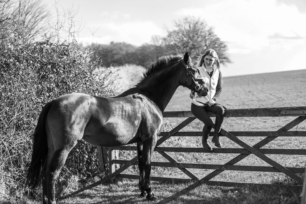 Black and white photo of a woman sitting on a wooden fence beside a horse in a rural field setting.  Essex Equine Photography.