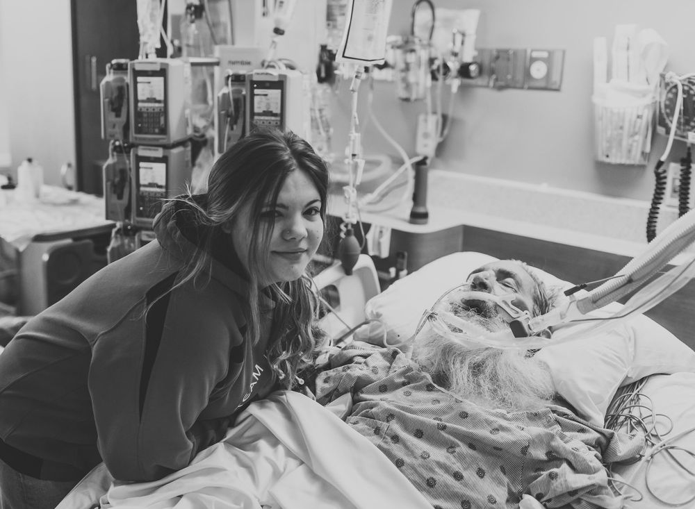 Medical patient lying in hospital bed with various medical equipment and monitors visible in the background.