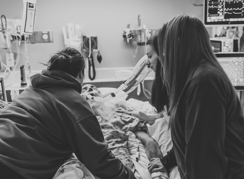 Black and white photo of visitors at a hospital bedside with medical equipment visible in the background.