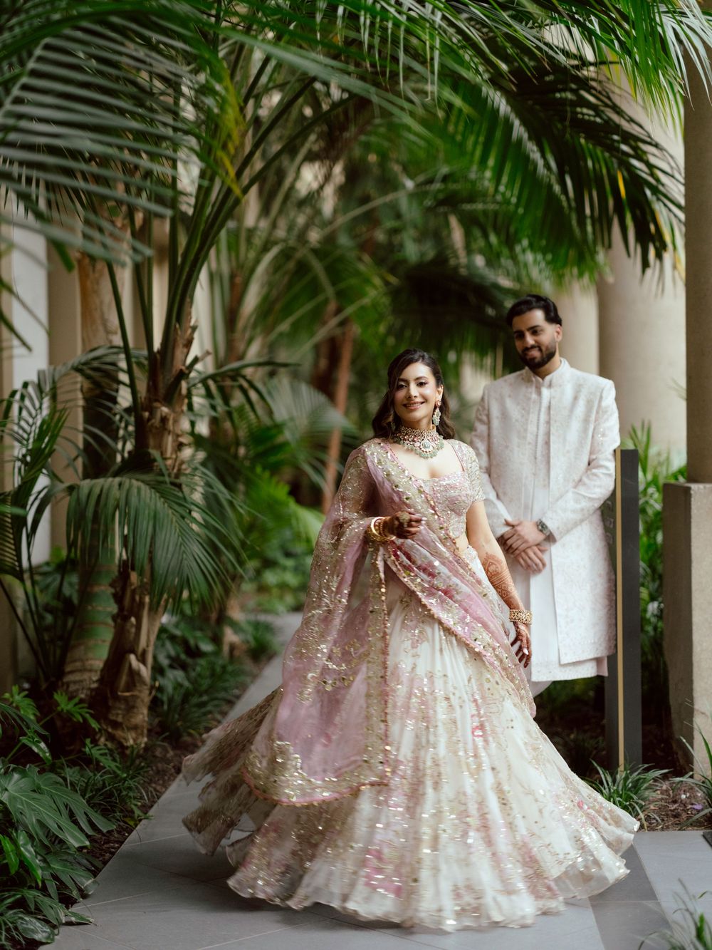 Couple in elegant attire poses together among lush palm trees in an outdoor courtyard setting.