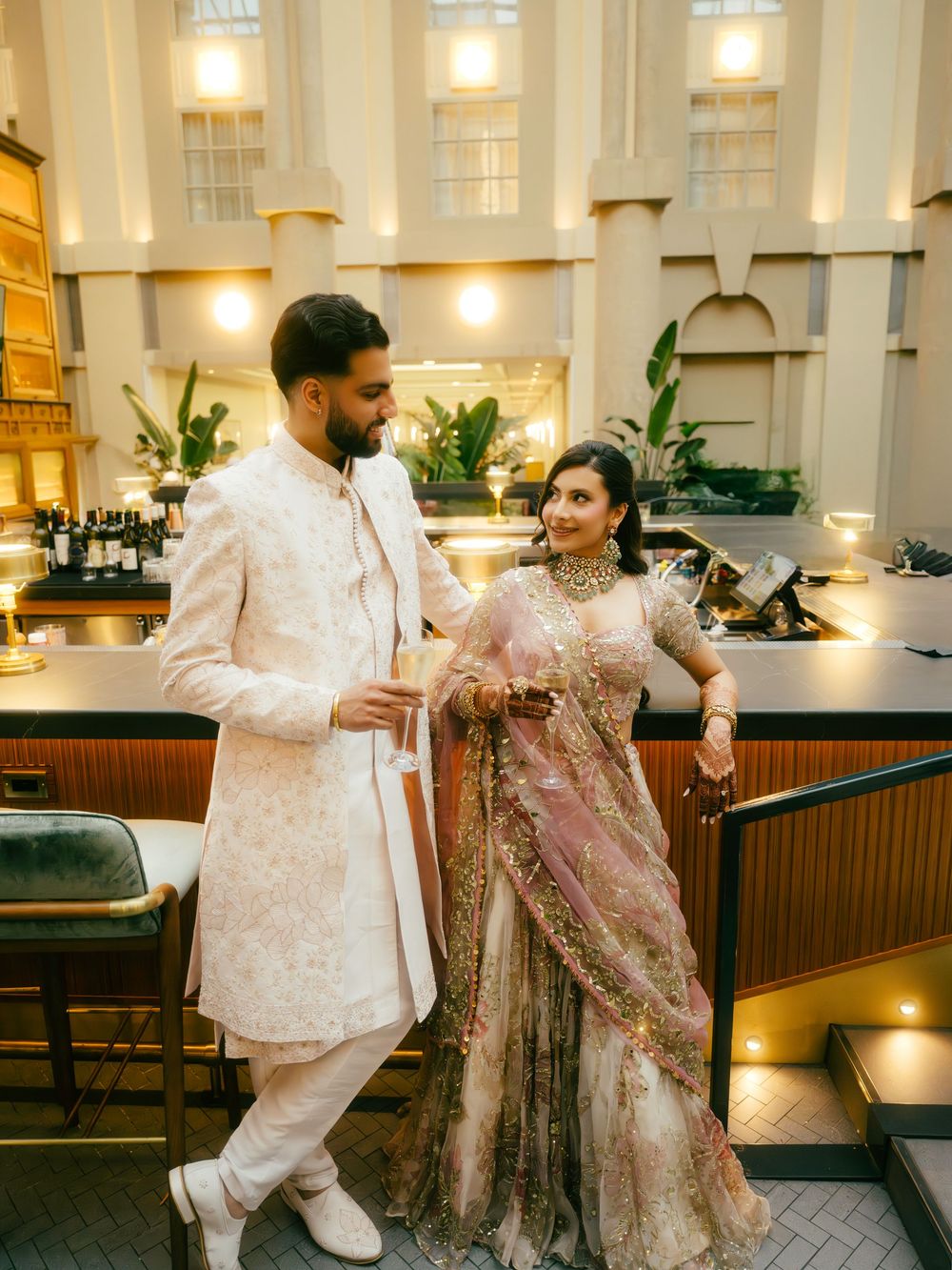 Couple in elegant South Asian wedding attire dancing playfully in an upscale indoor venue with warm lighting.