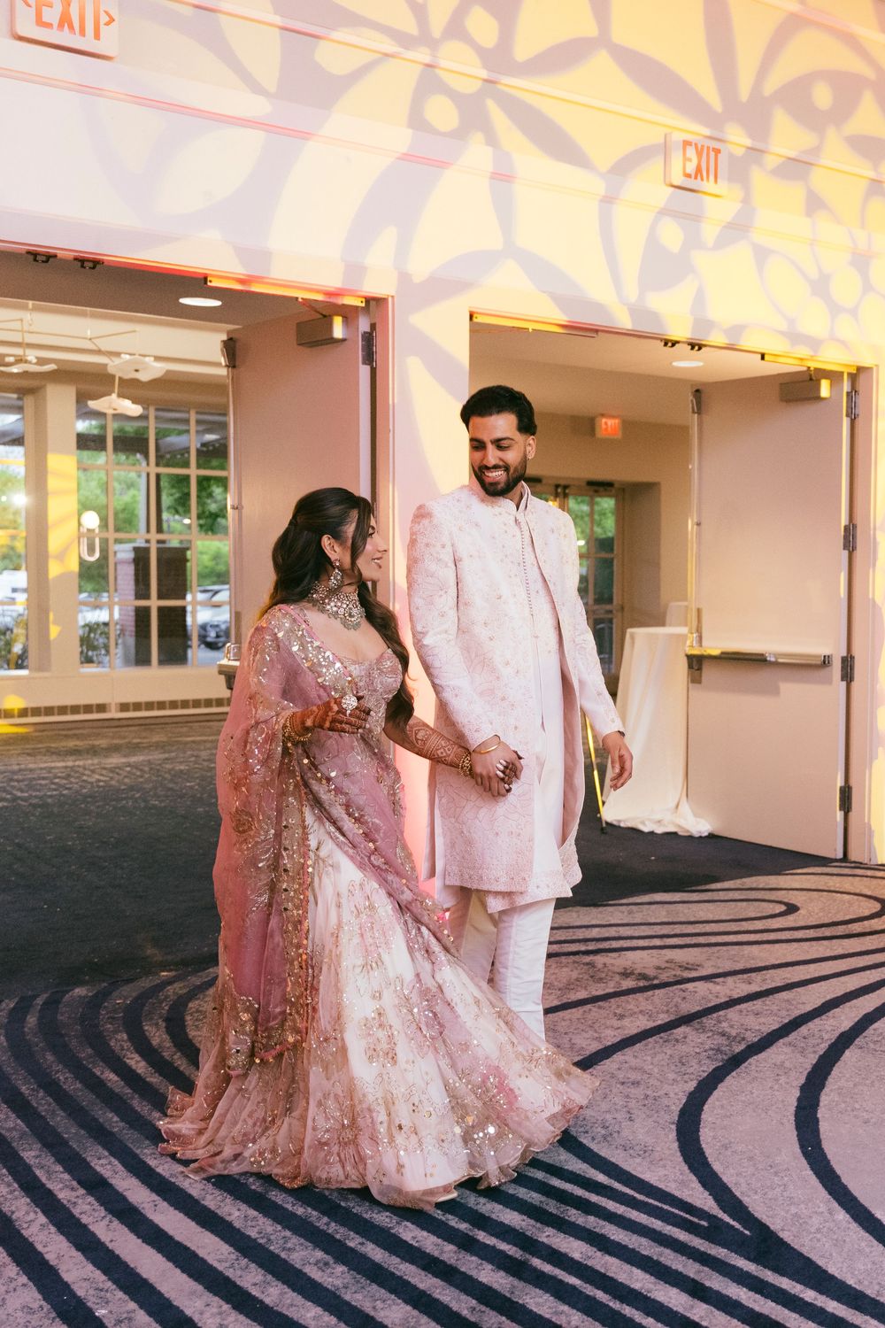 A couple in traditional South Asian wedding attire pose on a patterned carpet in an elegant indoor venue.