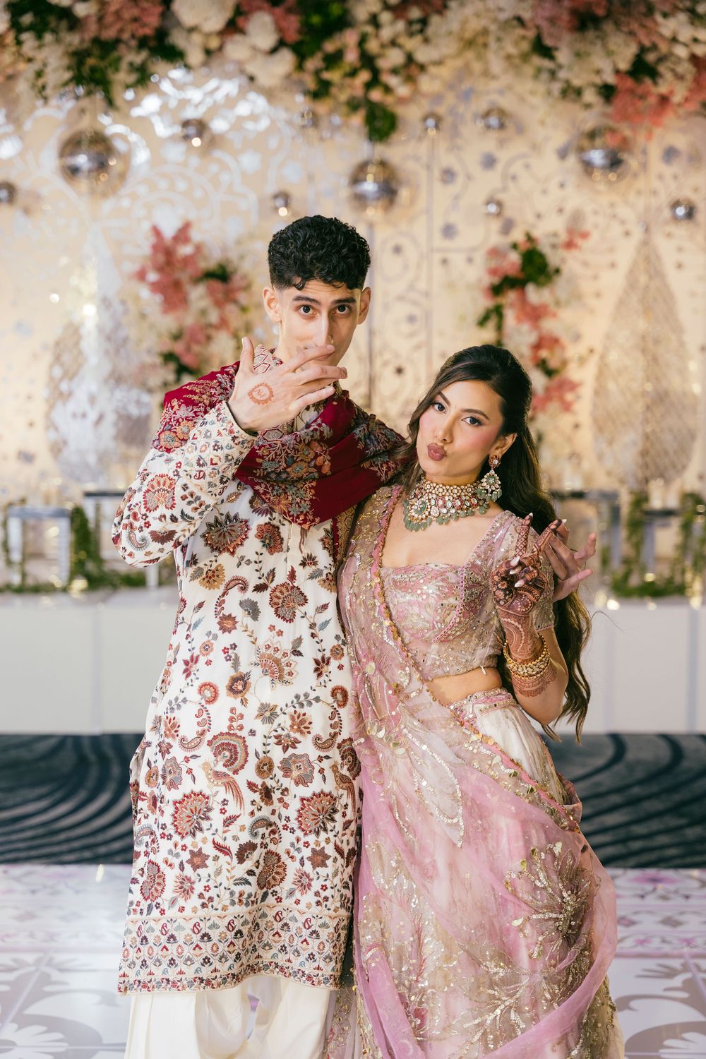 Couple in ornate pink and white wedding attire pose against a floral backdrop.