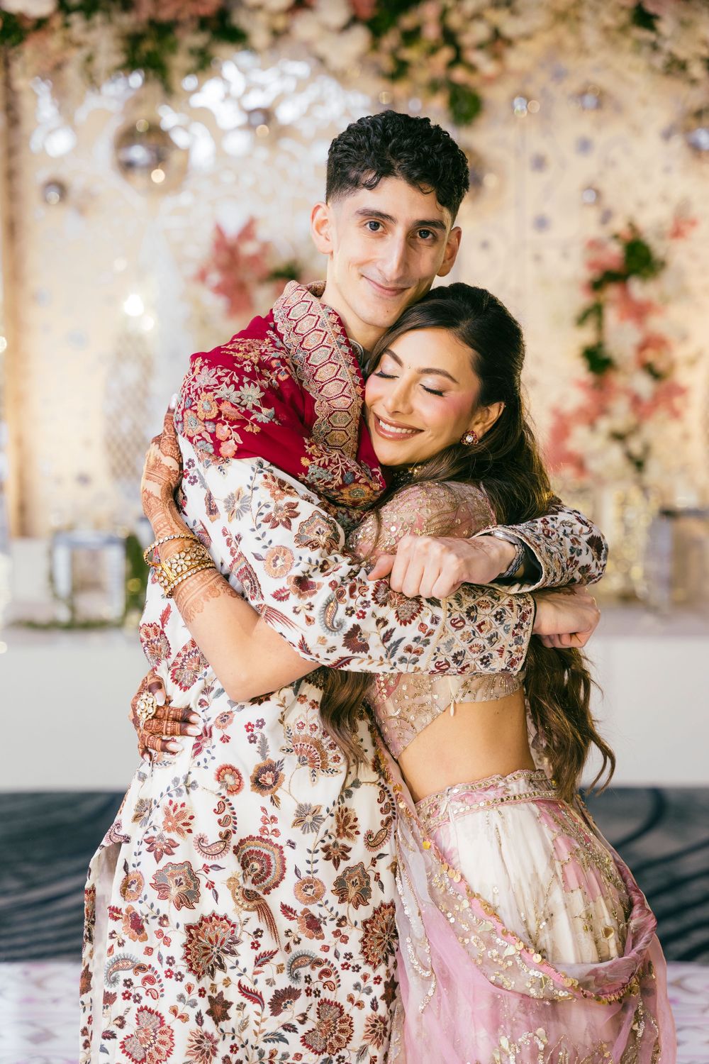 Couple embraces in traditional South Asian wedding attire with pink and red embroidered outfits against floral backdrop.