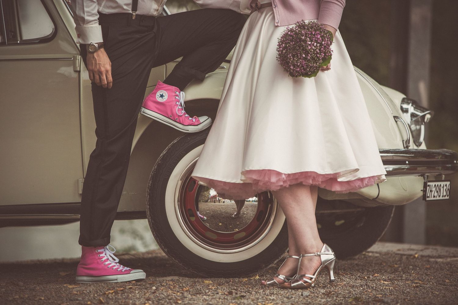 Couple in retro style leaning against vintage car, bride in white dress and groom in pink Converse.