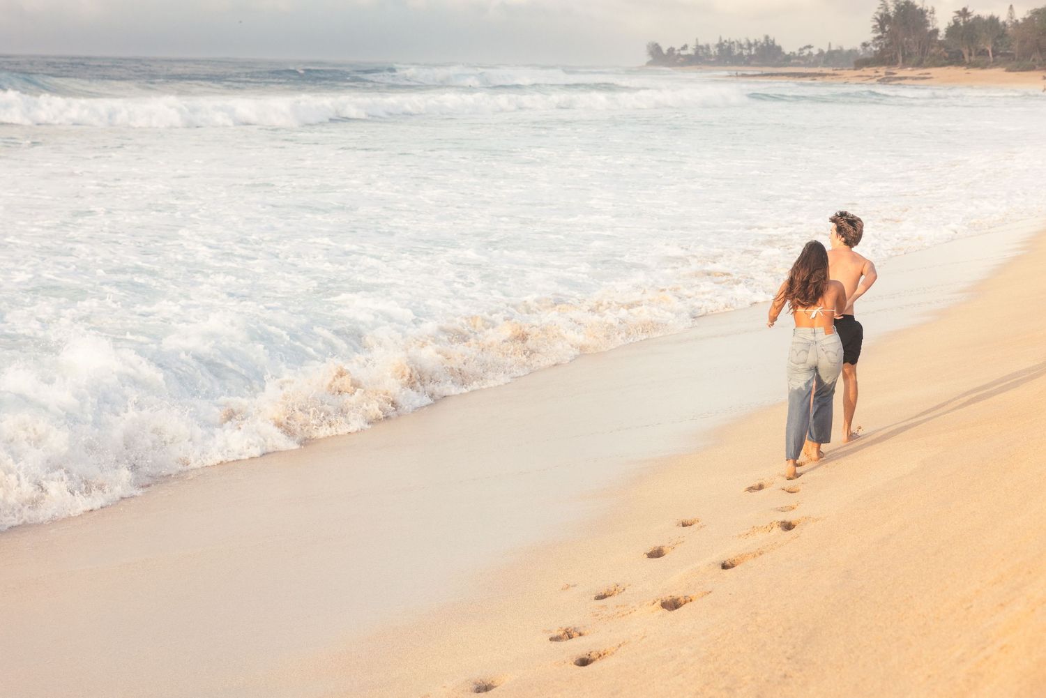 A couple walks along a sandy beach at sunset while ocean waves crash on the shore.