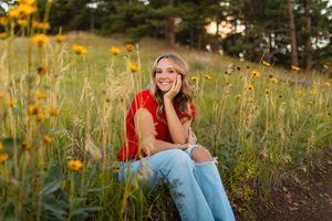 a girl sits in ripped jeans and red sweater among yellow wildflowers in a sunny meadow at golden hour for her aurora senior photos