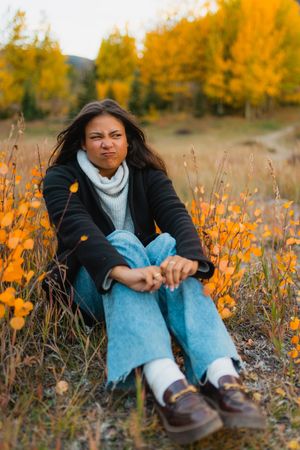 a high school girl sits in a field of golden wildflowers wearing light blue jeans, a black jacket and brown shoes on an autumn day.