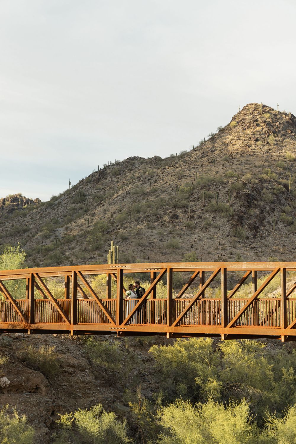 A rustic orange pedestrian bridge spans across a desert landscape with mountains and desert vegetation in the background.