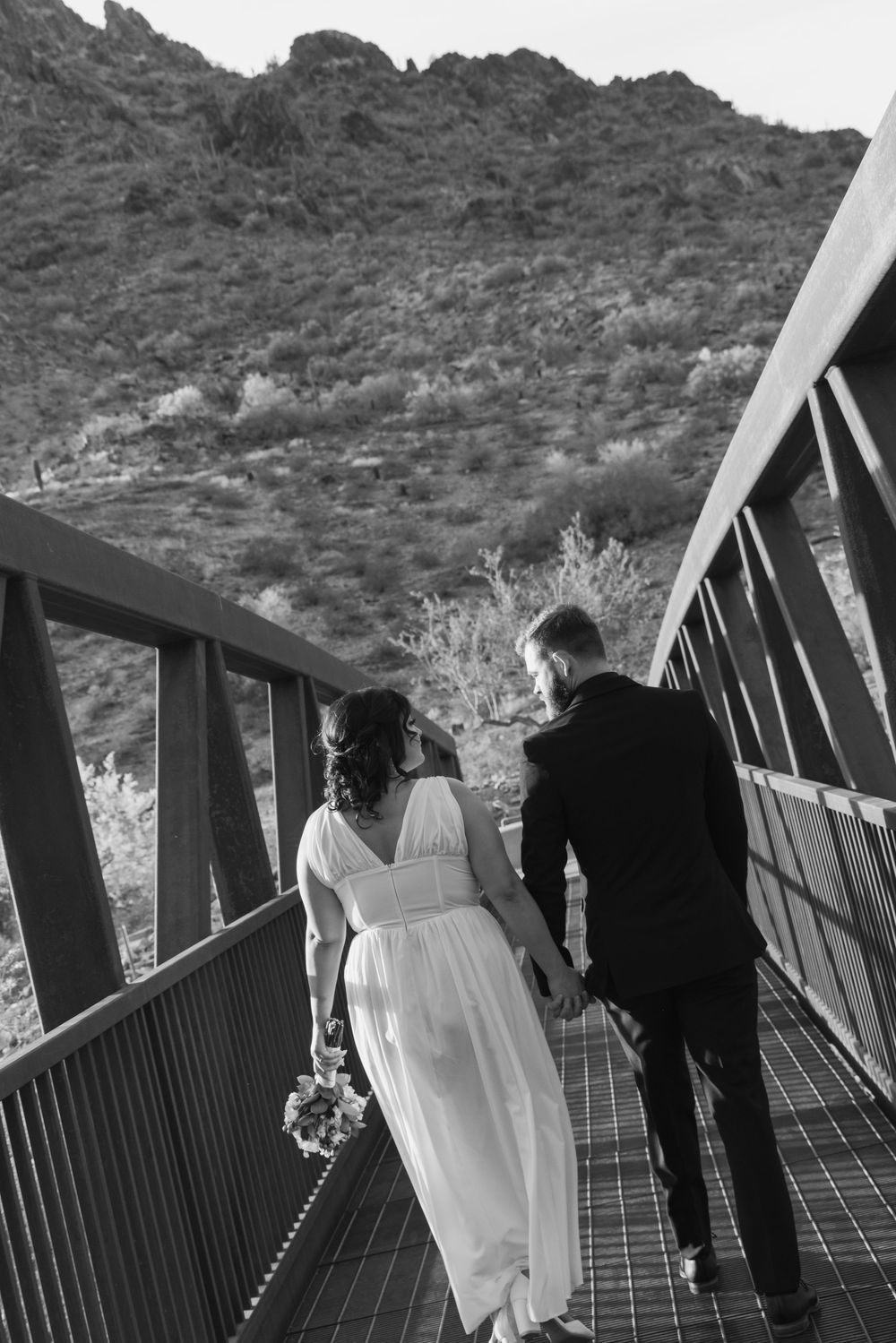 A couple walking across a metal bridge with desert mountains in the background in black and white photography.