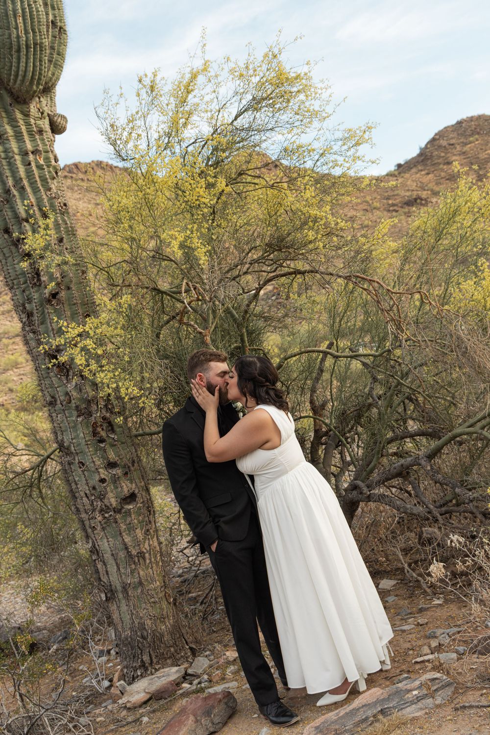 A couple in wedding attire embrace near a saguaro cactus in a desert landscape with mountains in the background.