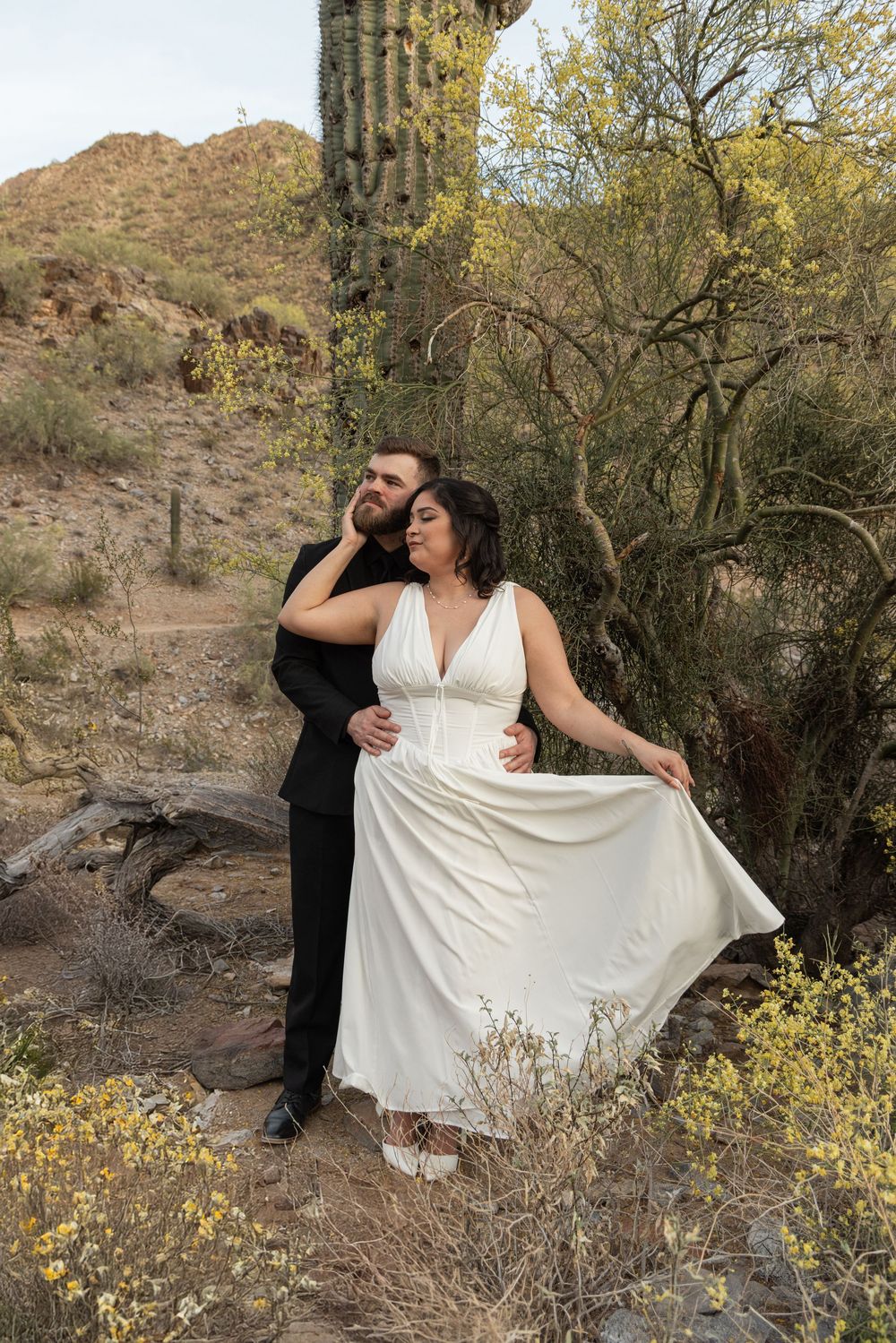 A couple poses together in the desert with a saguaro cactus and mountains in the background during sunset.