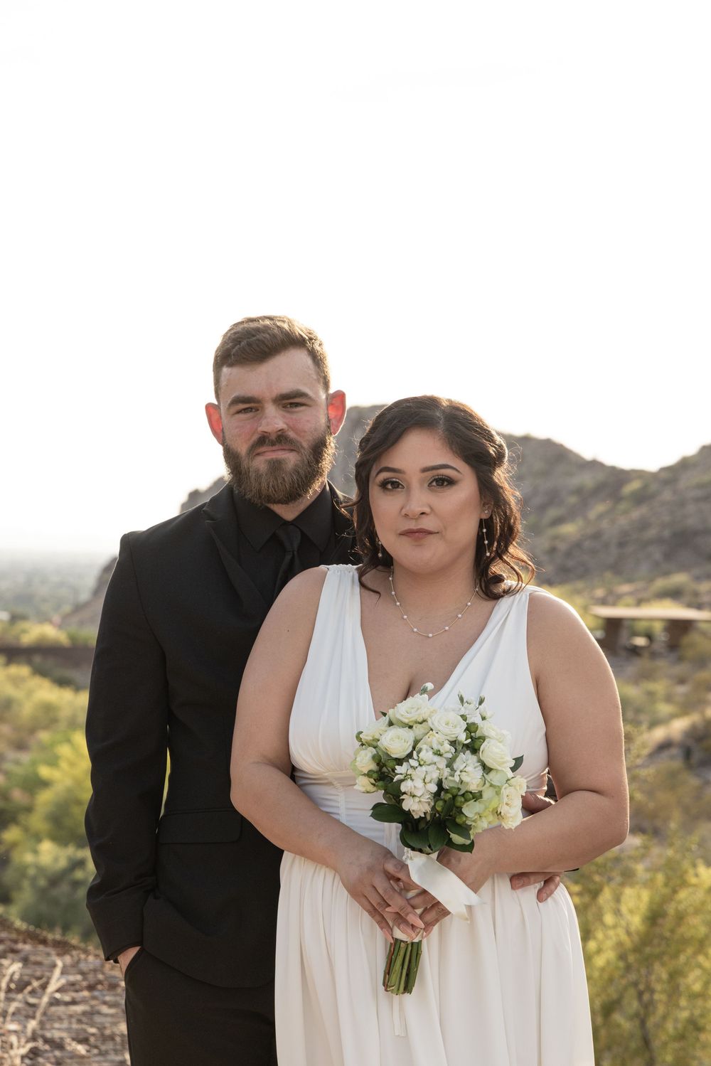 A couple in wedding attire poses together in the desert with mountains visible in the background.