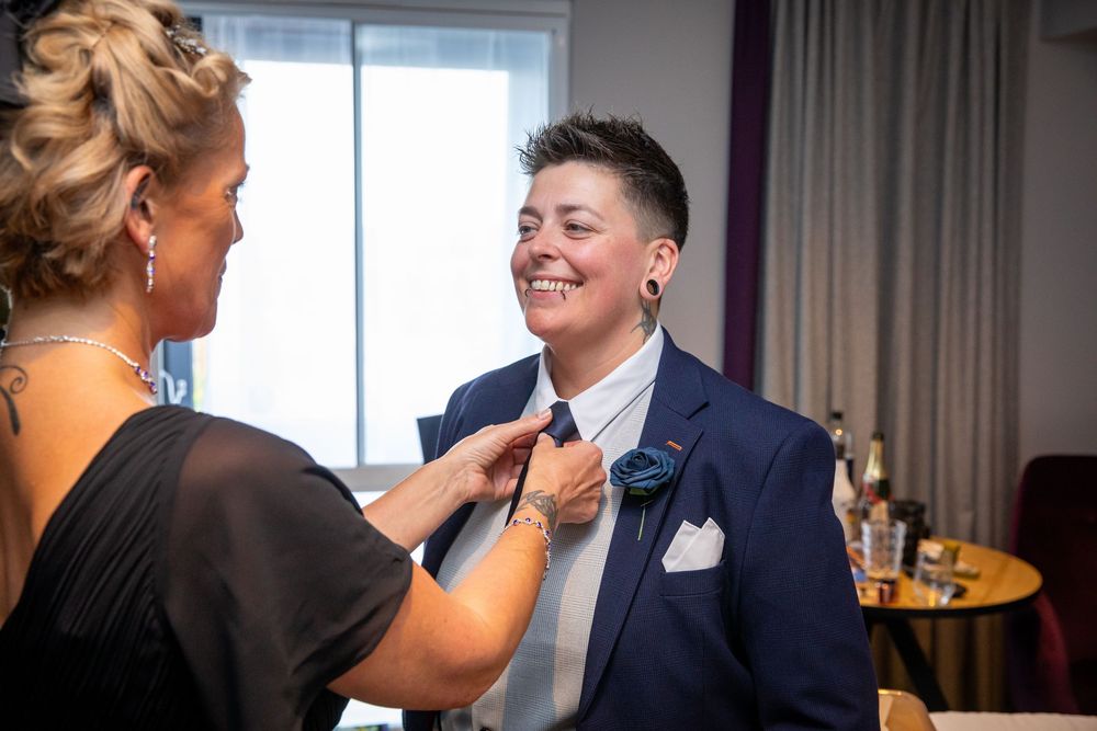 Woman adjusts tie of short-haired partner in navy suit with boutonniere before wedding ceremony.