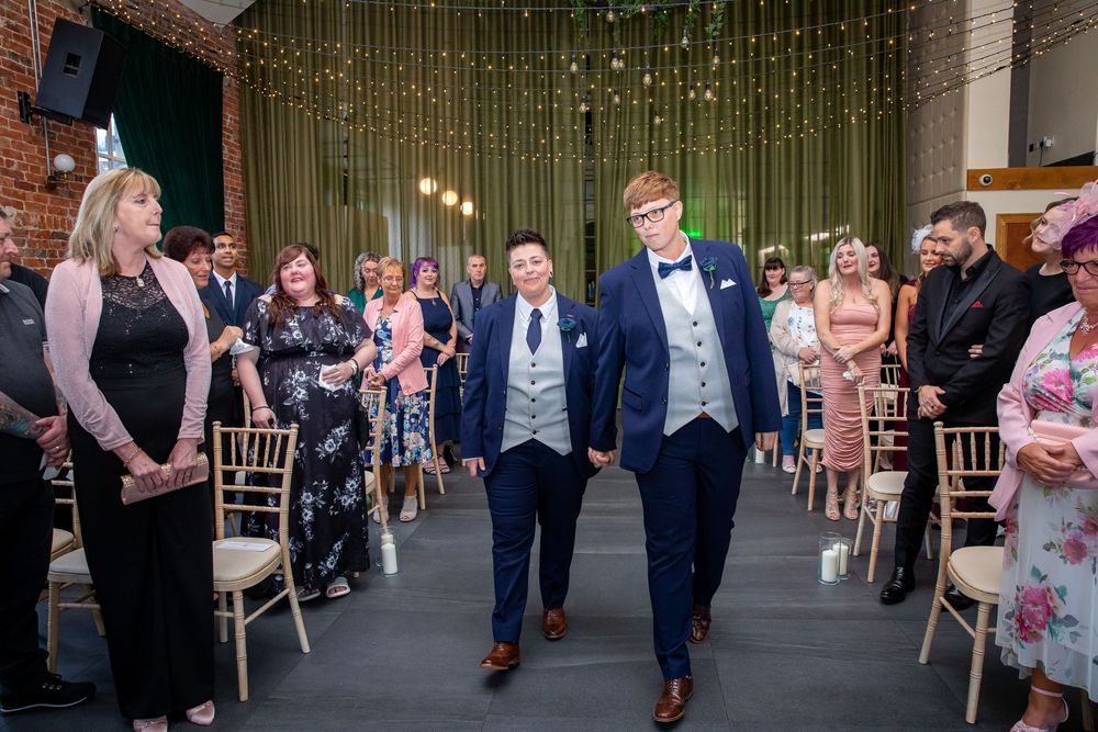 Two grooms in navy suits walk hand in hand down aisle as guests smile during wedding ceremony.