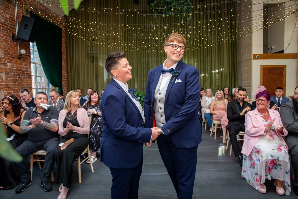 Two grooms in matching navy suits hold hands and smile during their wedding ceremony in a decorated venue.