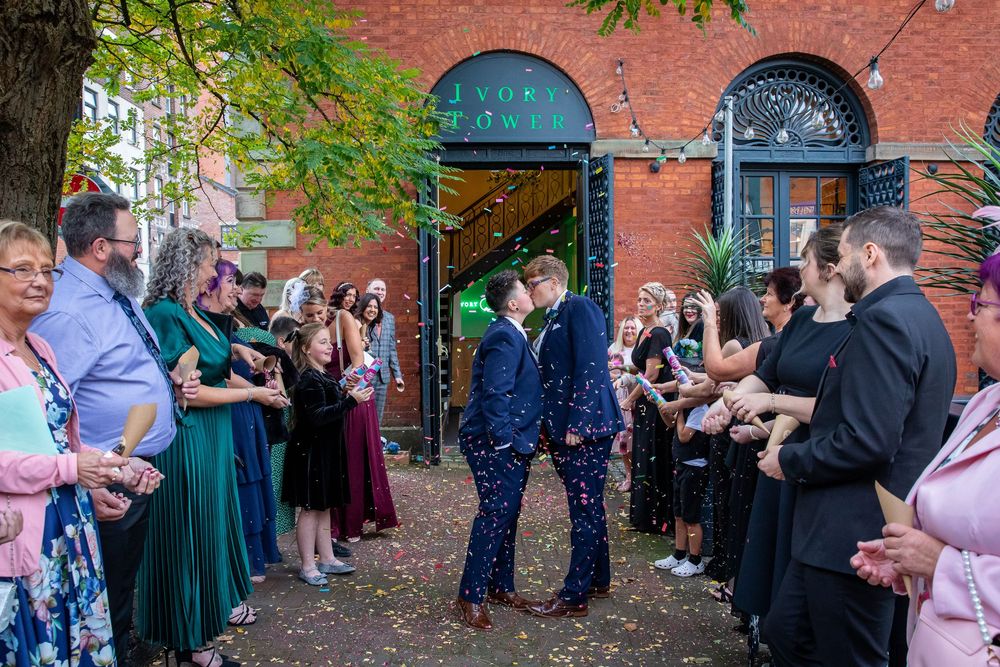 Newlywed couple exits through confetti shower surrounded by cheering guests outside a brick building.
