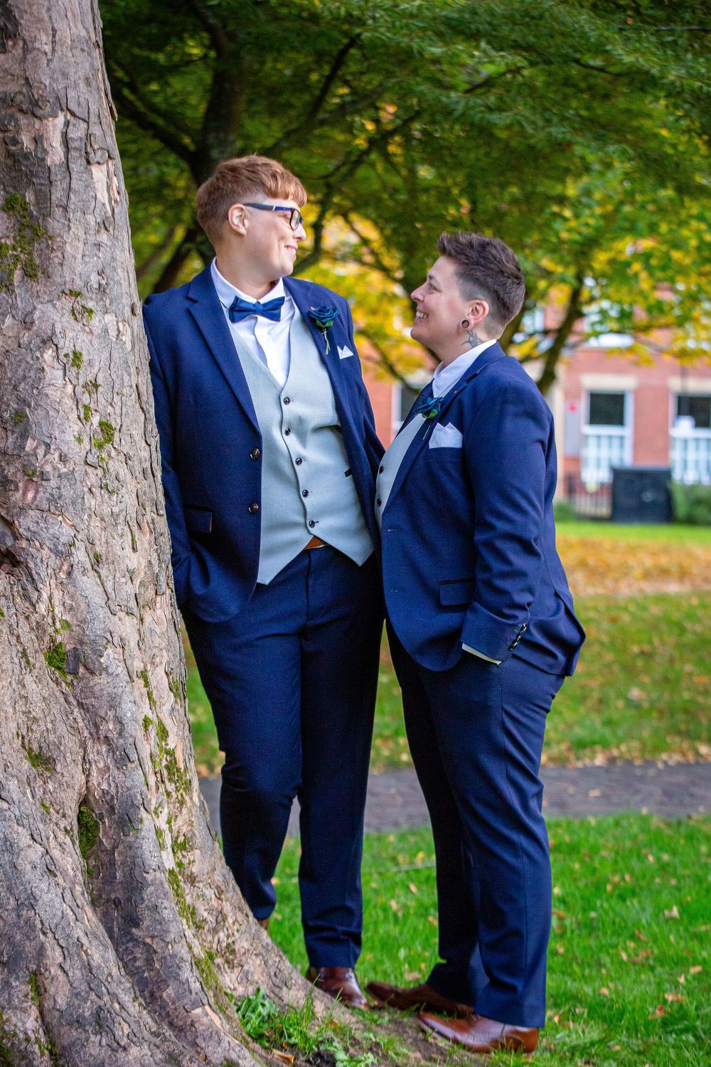 Two grooms in navy suits share a romantic moment leaning against a tree in a park setting.
