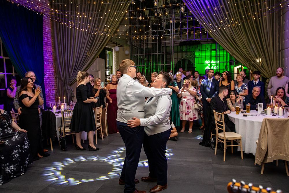 Two grooms share their first dance surrounded by guests in a beautifully lit wedding reception venue.
