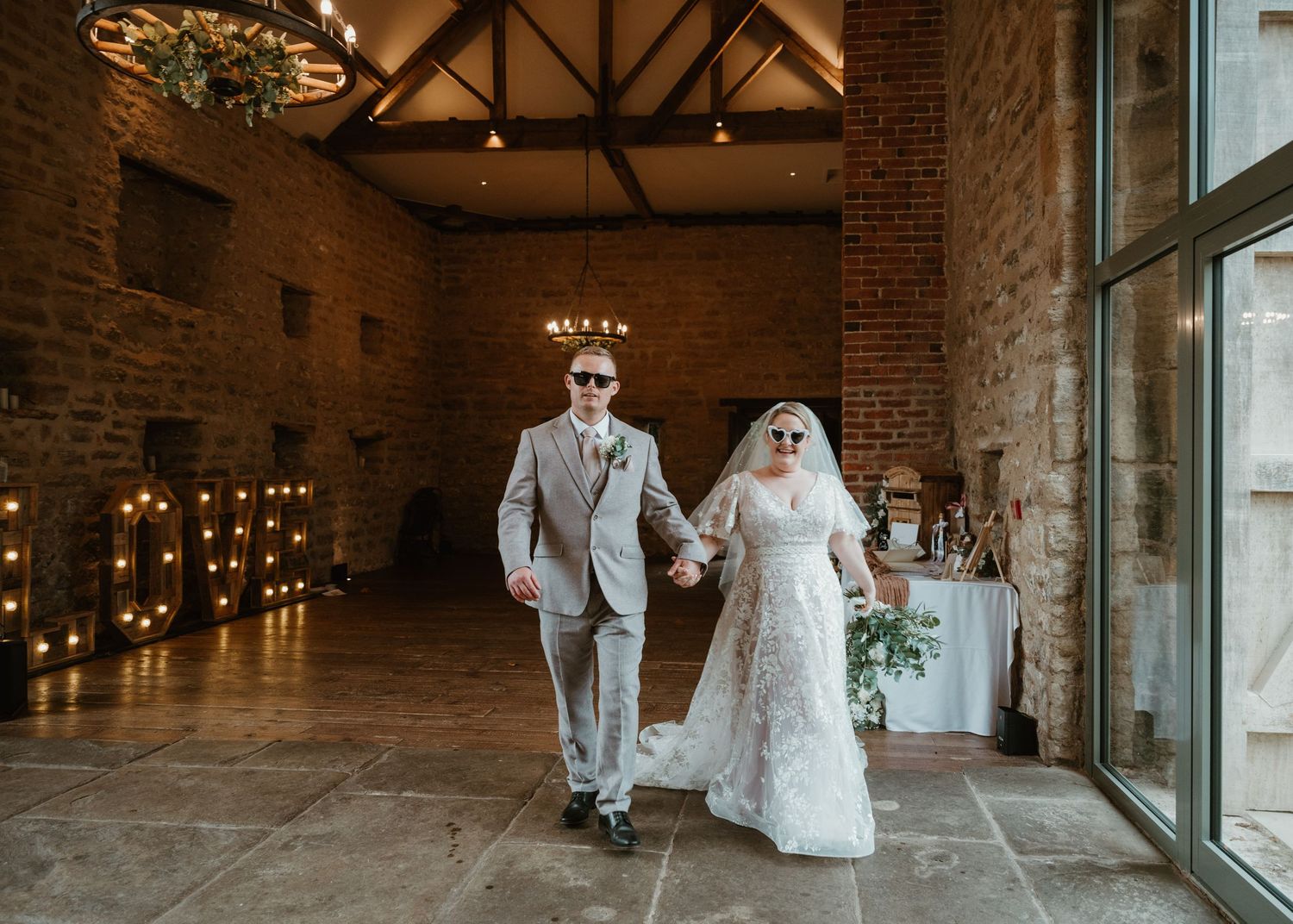 Wedding couple walks through a rustic brick venue with exposed beams and large windows creating a romantic atmosphere at Hooton pagnell hall