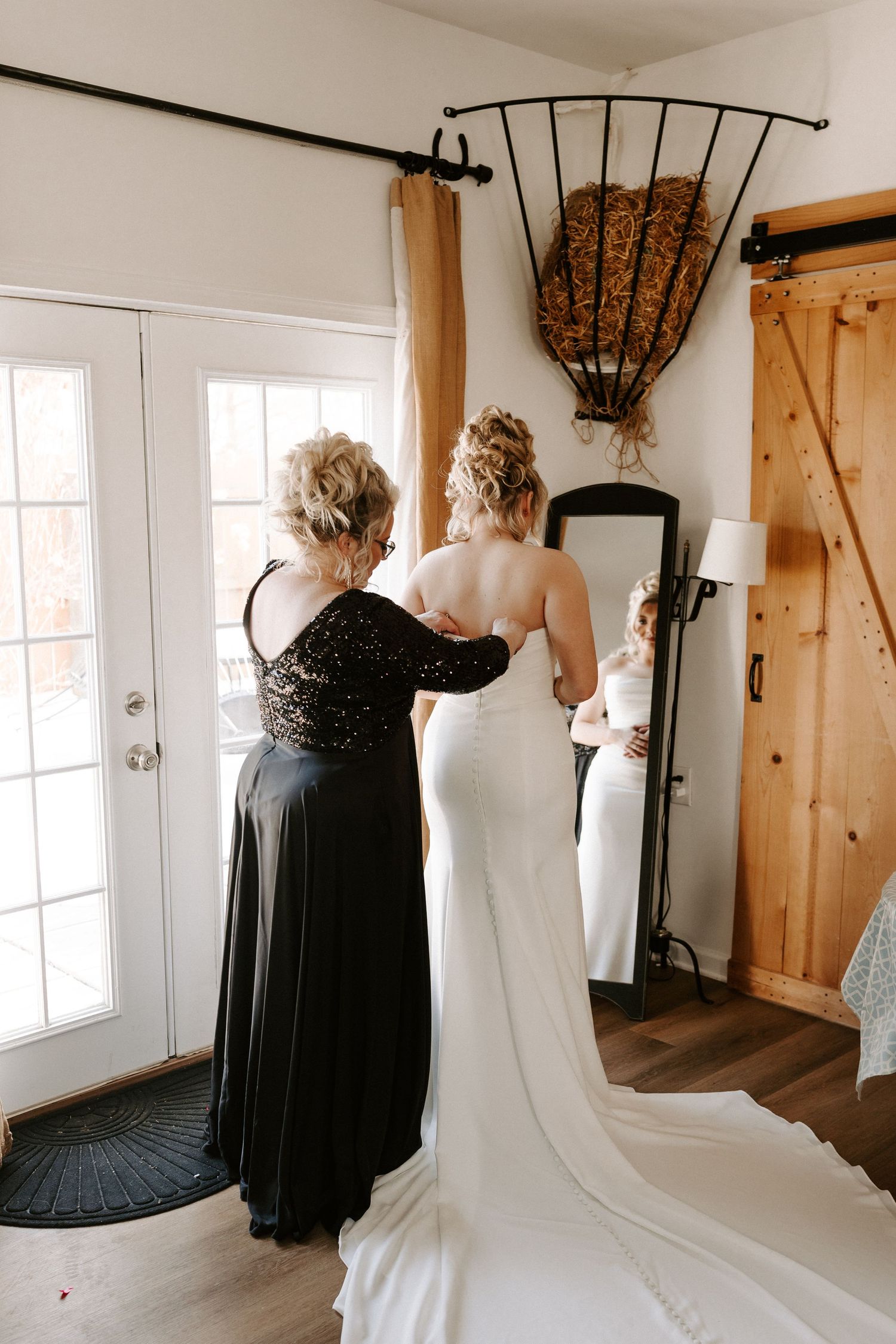 Mother helping bride button white wedding gown in rustic barn venue getting ready room.
