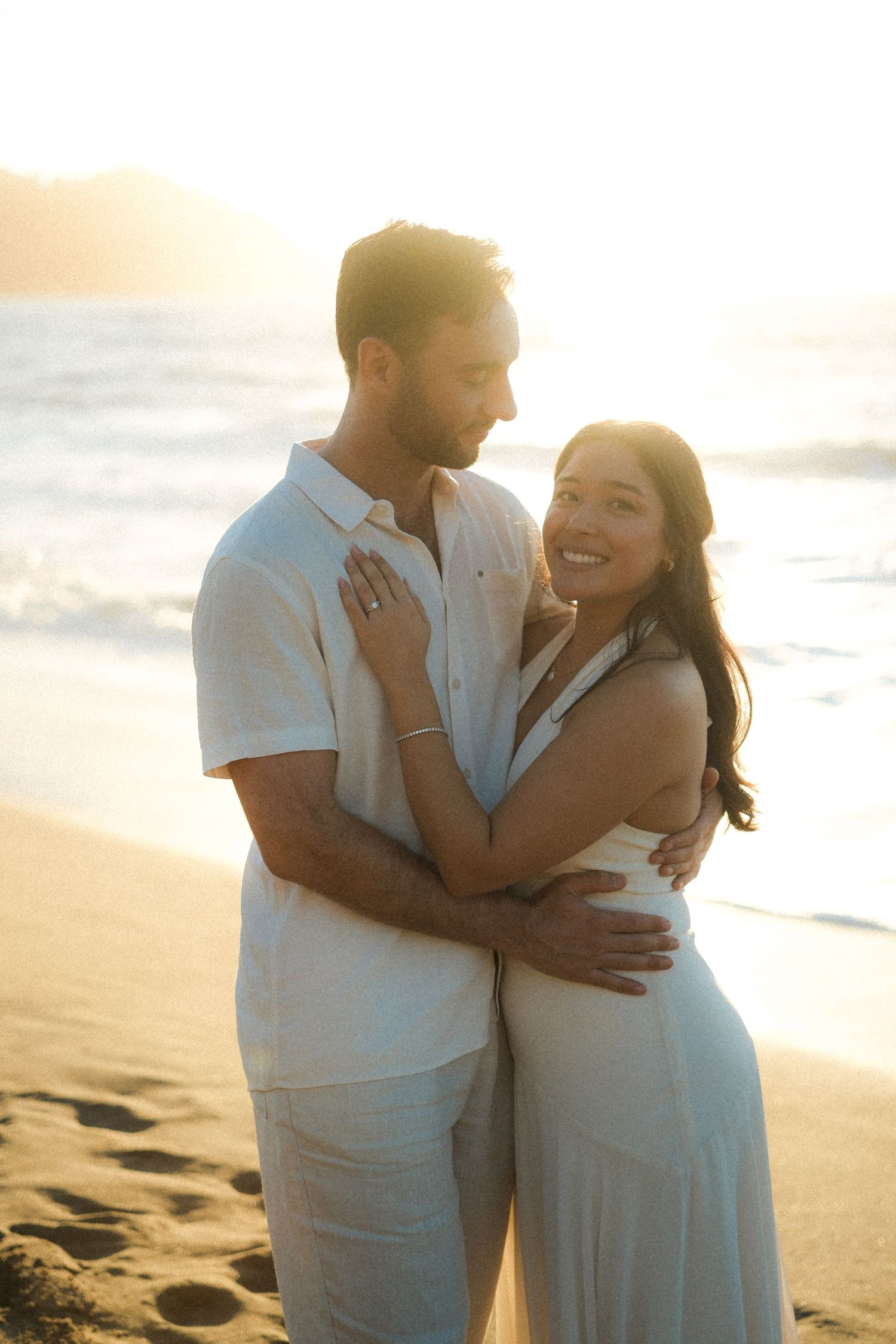 Romantic couple embraces on beach at sunset wearing white outfits with golden light in background.