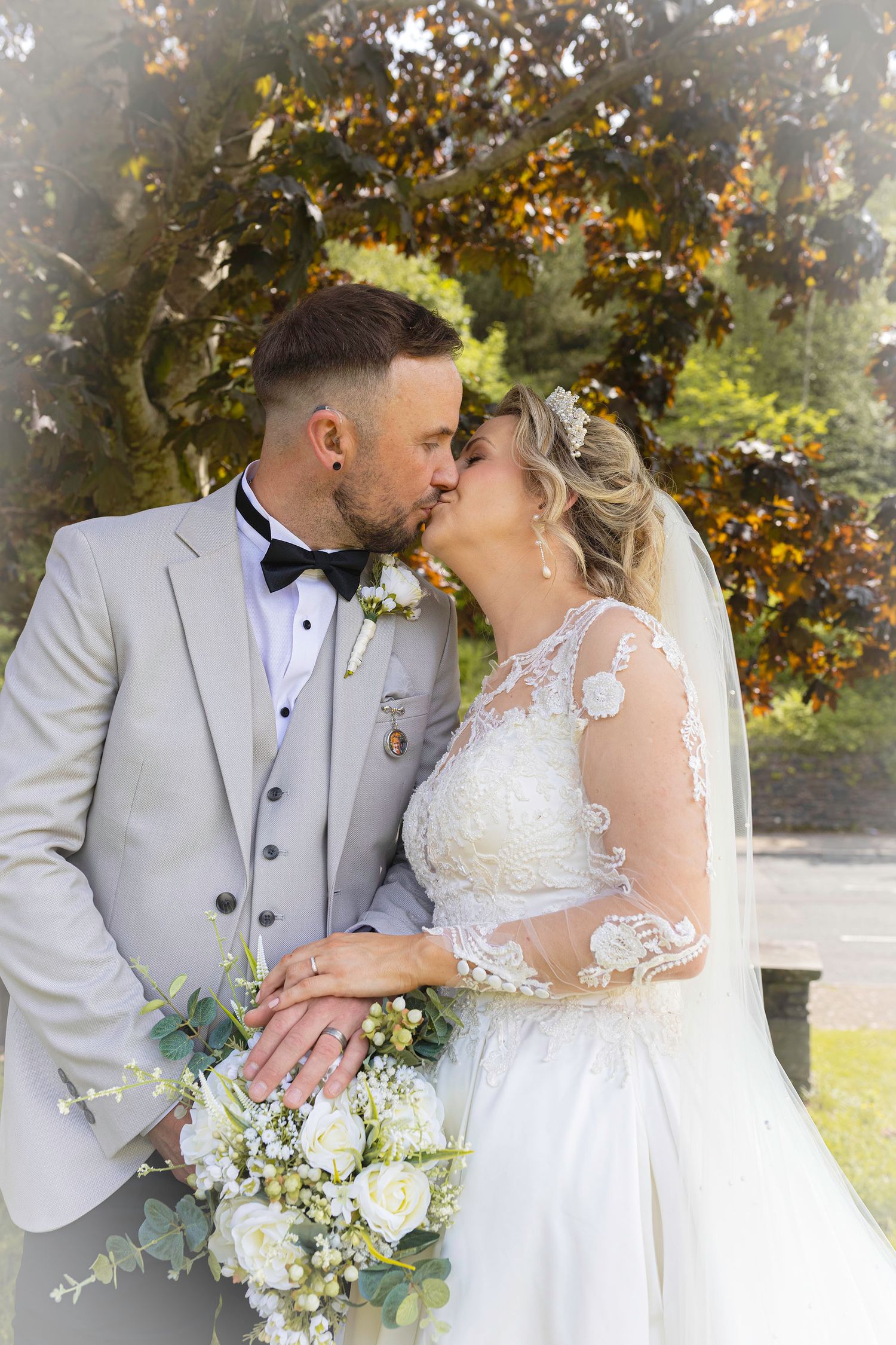 Bride and groom sharing a romantic kiss outdoors, holding a white floral bouquet with autumn trees in the background.