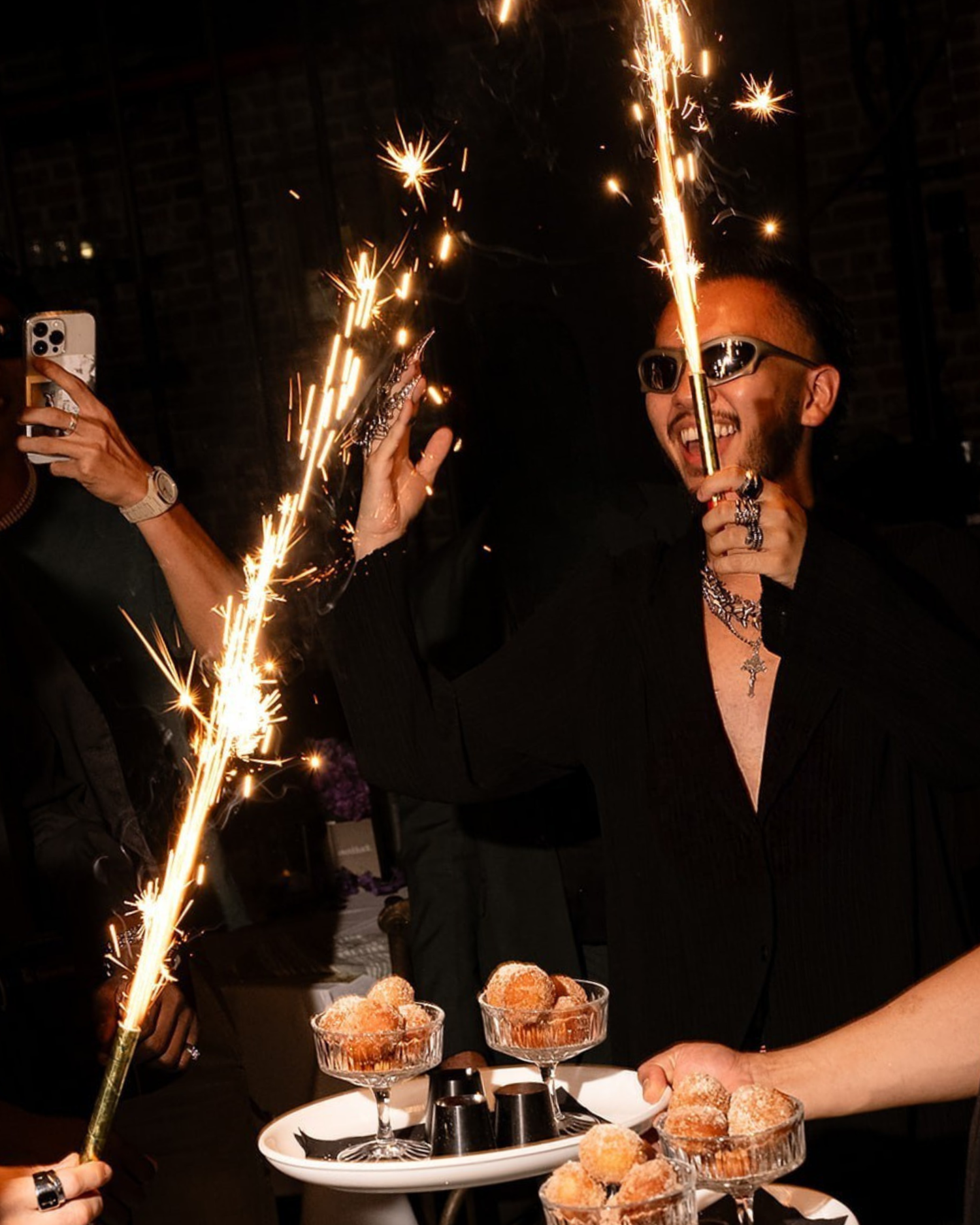 Tattooed man in black holding sparklers over desserts at a celebratory nighttime event.