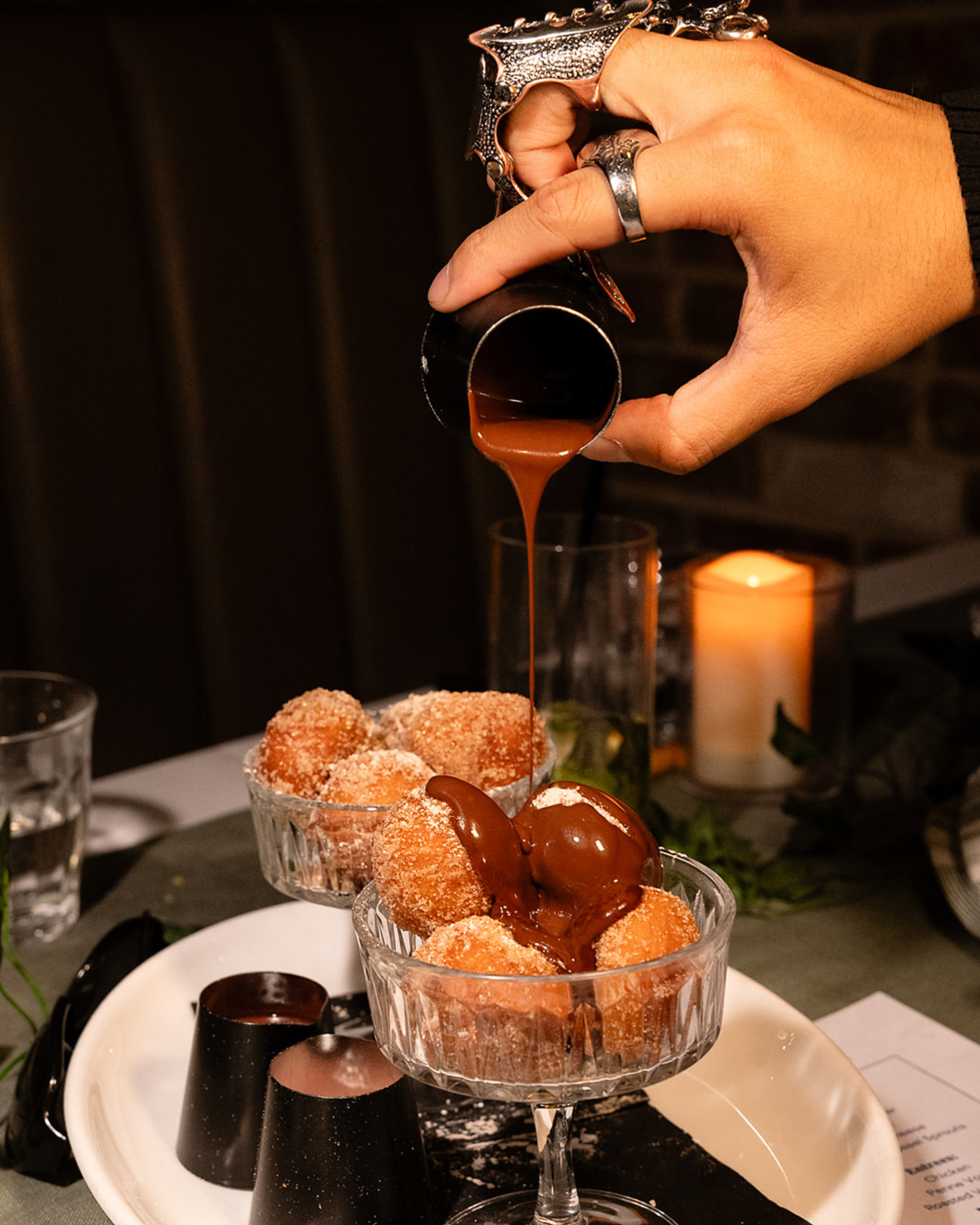 Hand pouring caramel sauce over sugar-dusted donut holes in a crystal bowl by candlelight.