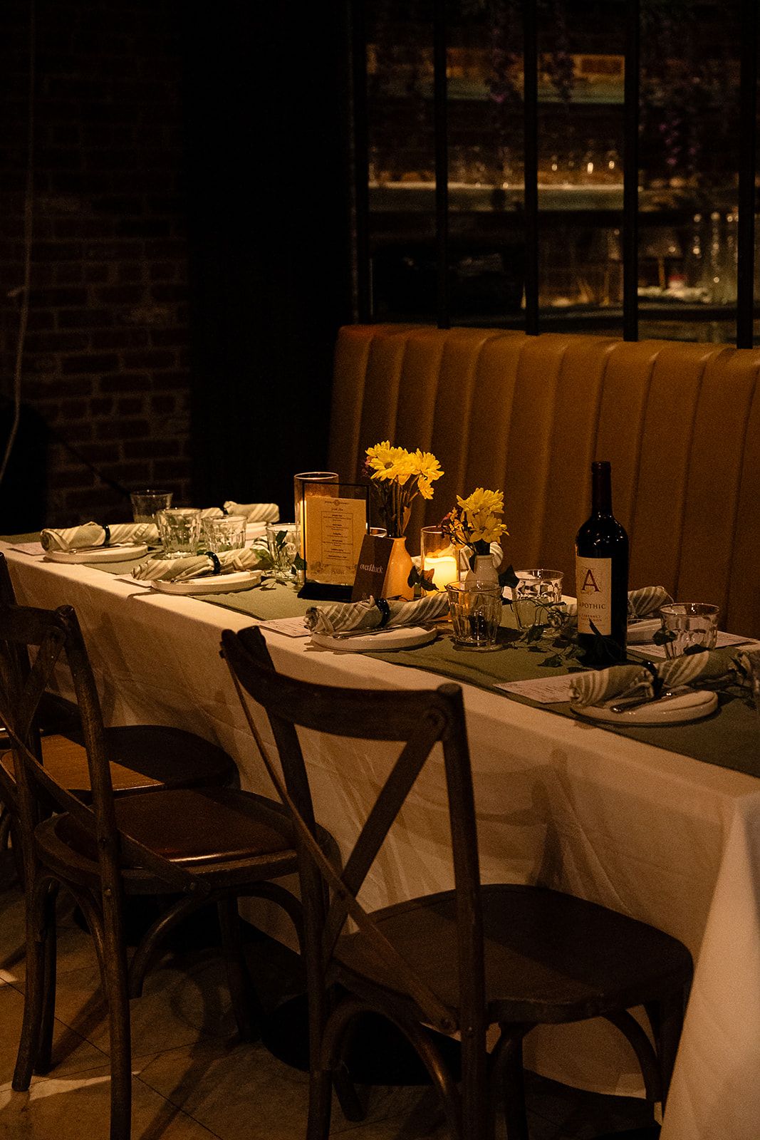 Elegantly set long dining table with candles, yellow flowers, and wine in a warmly lit restaurant.
