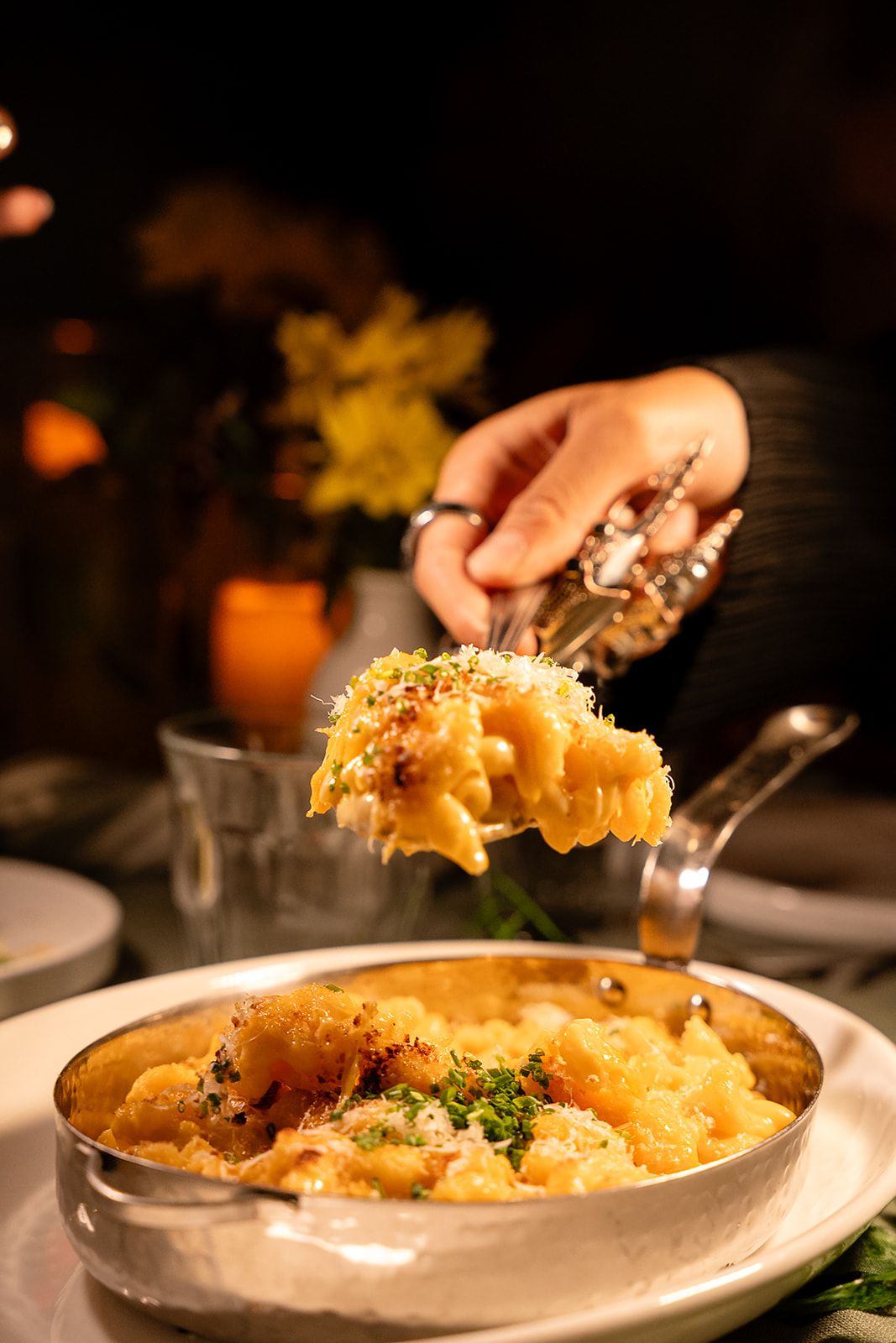 A hand scooping creamy baked mac and cheese from a silver pan in a dimly lit restaurant.