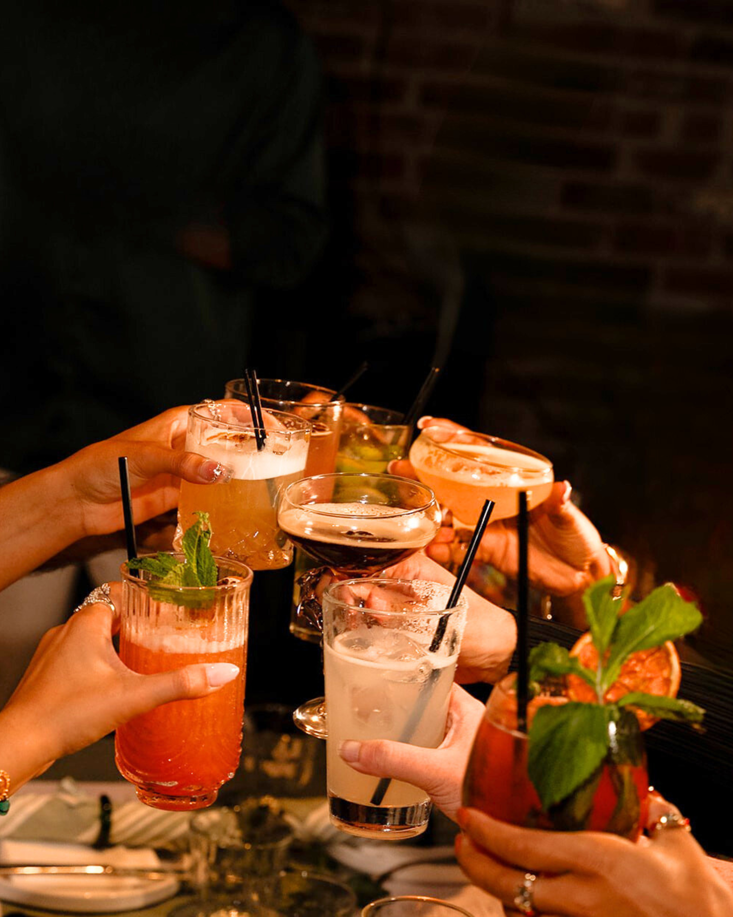 Group of friends toasting with colorful cocktails in a dimly lit brick-wall bar setting.