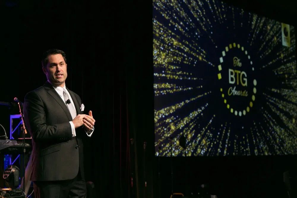 A man in a suit speaks on stage at the BTG Awards ceremony, with a glowing star-burst logo behind him at the Dallas Renaissance Hotel